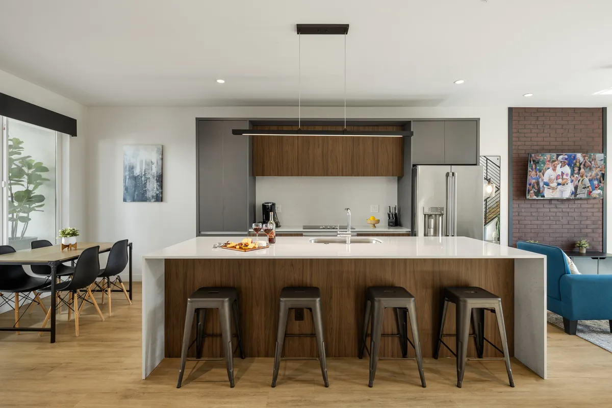A modern kitchen and dining area with a spacious kitchen island, sleek stools, contemporary lighting, and a dining table situated nearby. The kitchen is equipped with high-end appliances and receives abundant natural light, creating an inviting space for group cooking and shared meals.