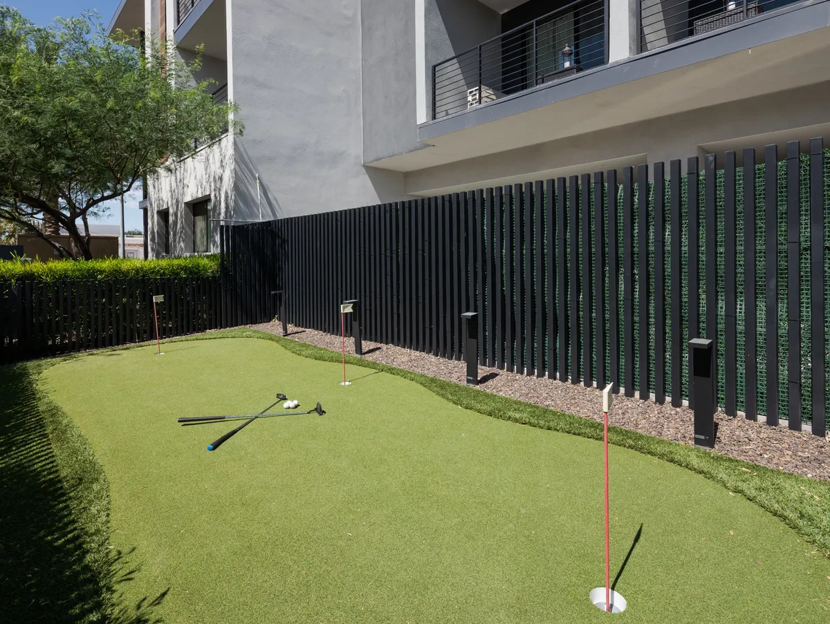 A modern putting green bordered by attractive landscaping and contemporary architecture. Golf clubs and casual seating invite friendly play and outdoor relaxation.