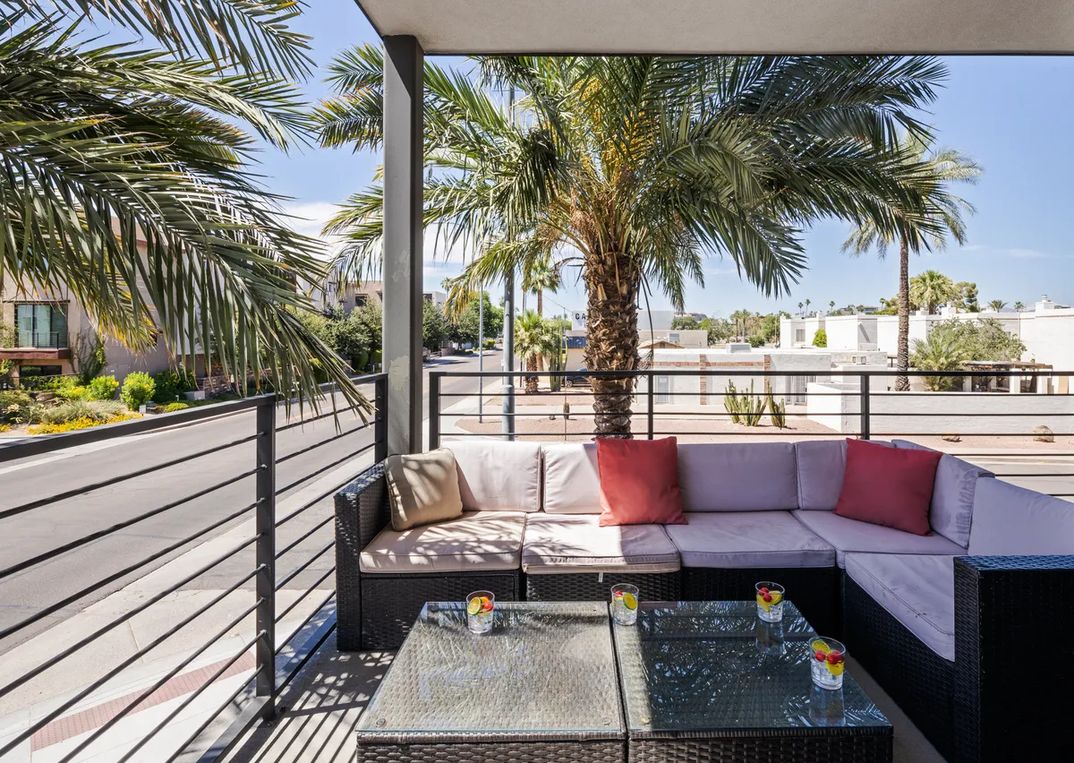 The image captures an inviting outdoor seating area shaded by swaying palm trees, featuring a stylish glass coffee table surrounded by plush, decorative cushions. The scene is bathed in warm Arizona sunlight, suggesting a perfect spot for relaxing with a drink, socializing with friends, or starting the morning with coffee. The lush greenery and elegant setup speak to a luxurious, resort-style ambiance right in the heart of Old Town Scottsdale.
