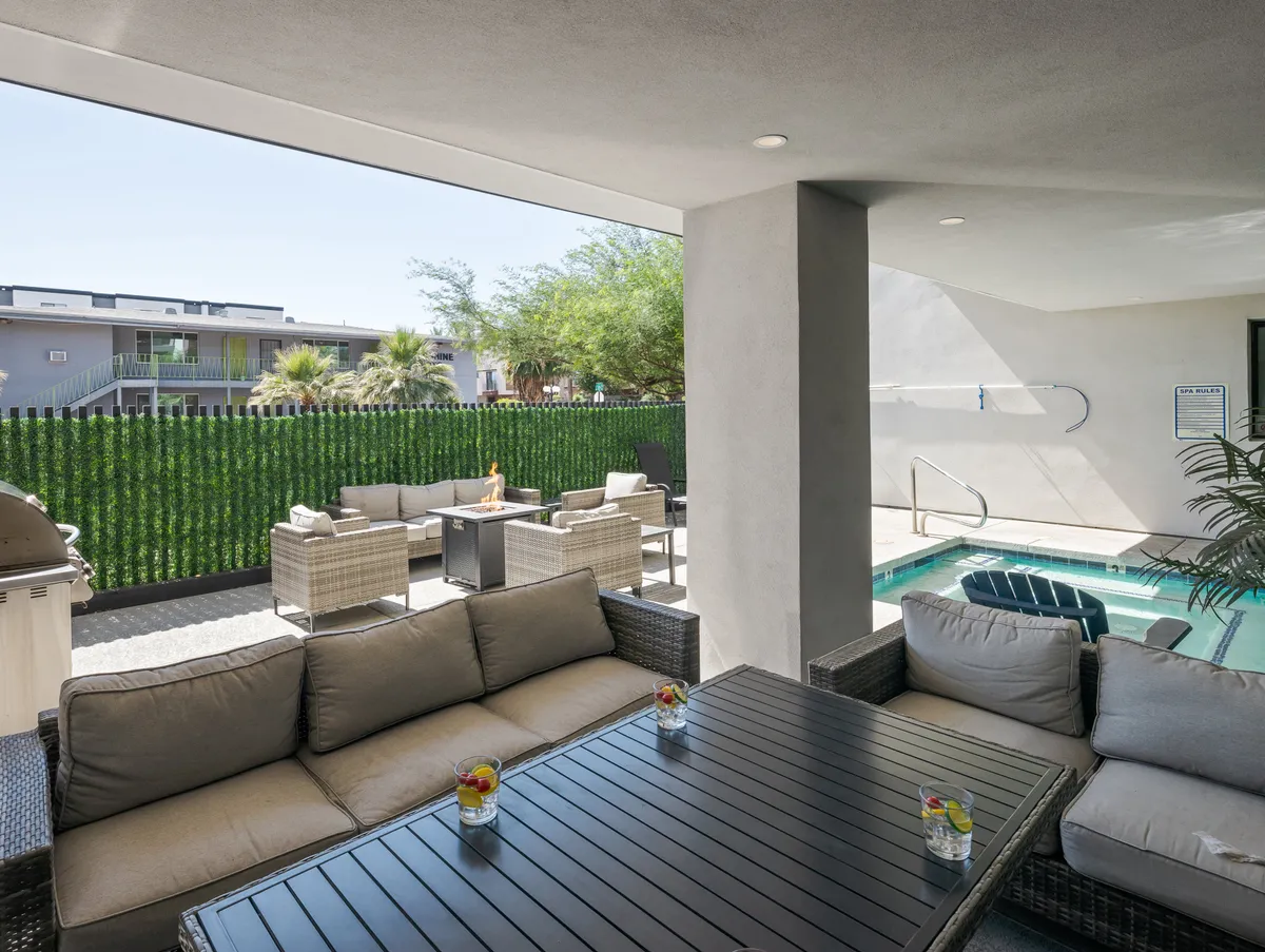 A lively exterior scene featuring a sparkling swimming pool, adjacent outdoor seating area, and a table set with drinks, all framed by lush greenery. The setting exudes a leisurely and social mood, ideal for sunbathing, outdoor meals, or group relaxation.