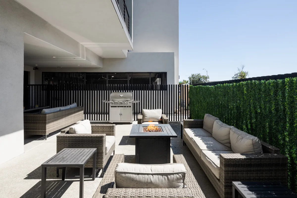 An inviting outdoor communal space with a cozy fire pit, comfortable seating arrangements, a BBQ grill, and lush greenery in the background. The area is perfect for group gatherings, outdoor dining, and sunset relaxation.
