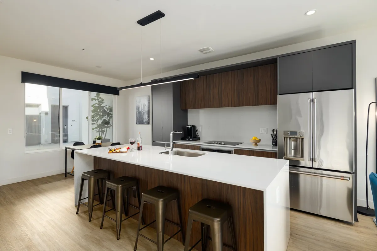 A bright, contemporary kitchen with a spacious island, stainless steel appliances, and bar stools, filled with natural light from the windows. The scene suggests a welcoming environment perfect for cooking, entertaining, and family gatherings.