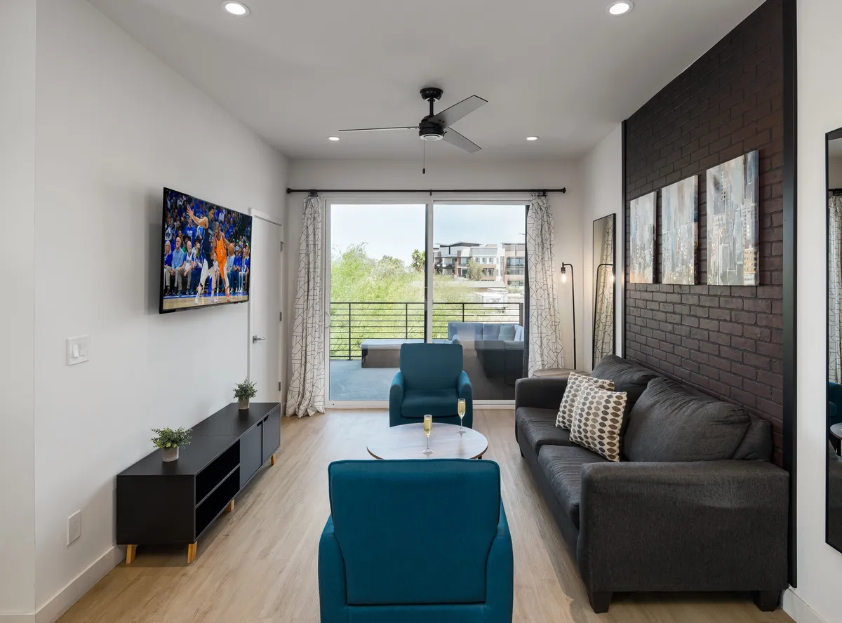 A bright, open living room featuring a sofa with patterned cushions, vibrant blue chairs, a television displaying a sports game, and large sliding glass doors leading out to a private balcony. Sunlight streams through, giving the space a cheerful, inviting atmosphere ideal for group gatherings, relaxation, and entertainment.