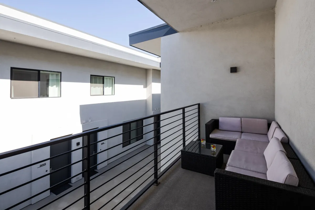 An exterior balcony scene showcasing an outdoor seating area with a small table set for drinks, surrounded by a casual atmosphere and views over the neighborhood. The space is ideal for intimate gatherings, watching the sunset, or enjoying a quiet coffee with friends and family.
