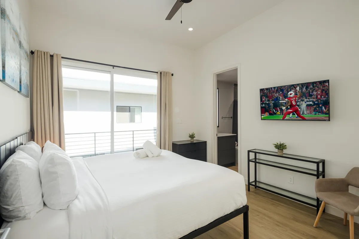 A cozy bedroom with crisp white linens on the bed, a television displaying a sports game, and a large window letting in natural light. The room feels comfortable and contemporary, ideal for both relaxation and leisure.