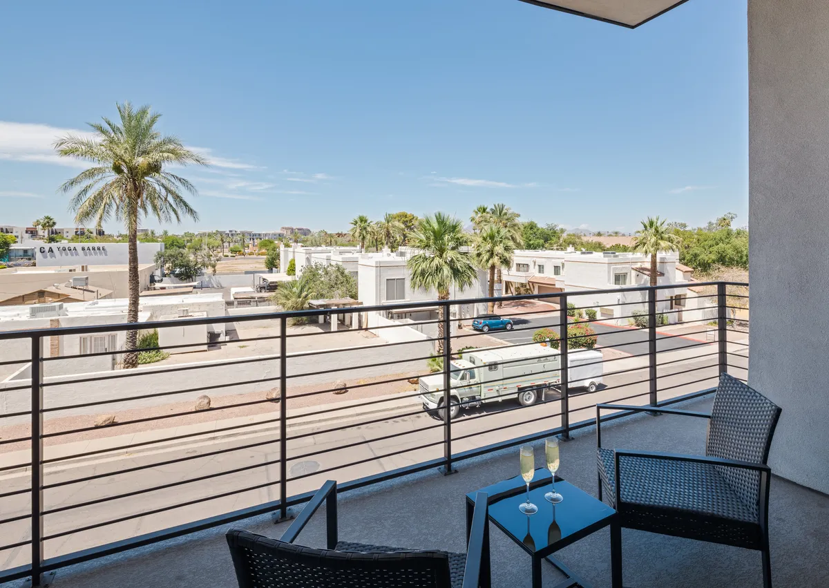 A tranquil balcony scene with chairs and a table, framed by palm trees and an urban landscape. The space is ideal for unwinding in the evening, enjoying a nightcap, or gathering with friends to reflect on the night’s adventures.