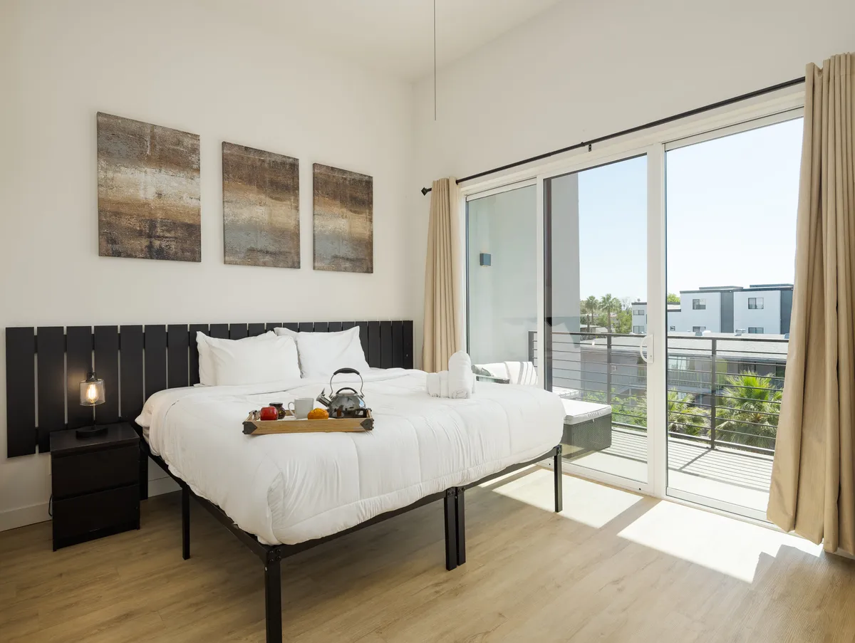 A serene bedroom with white linens, abstract wall art, and a sliding glass door leading to a balcony. The room is filled with natural light, exuding tranquility and plush comfort, inviting relaxation and quiet mornings.